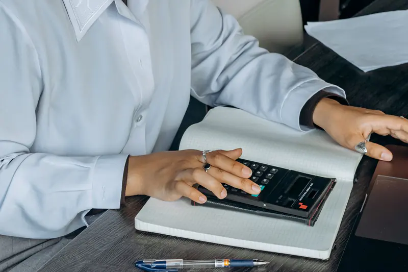 Mains d'une femme qui fait ses comptes avec une calculatrice, sur un carnet à côté d'un ordinateur portable